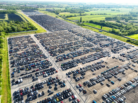 Aerial view of a massive car storage facility with thousands of vehicles parked in rows surrounded by green fields in Rivalta di Torino, Piedmont, Italy.