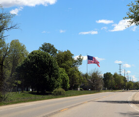 Fototapeta premium Asphalt road lined with green trees and an American flag under a blue sky