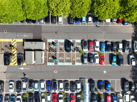 Aerial view of a parking lot with rows of cars and electric vehicle charging stations next to green trees in Turin, Piedmont, Italy.