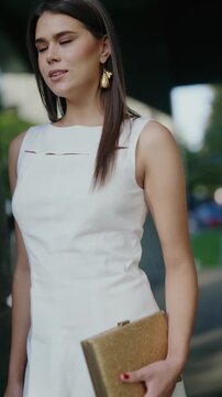 A stylish brunette woman in a white dress and gold clutch walks confidently outdoors. She embodies elegance and sophistication, perfect for lifestyle, fashion or beauty projects.