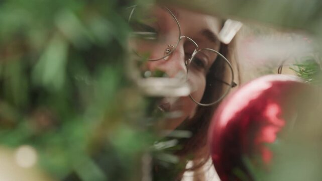 Young white woman designer inspecting ornament on lush evergreen tree with round glasses, hands arranging tinsel, focused expression under warm indoor light in cozy holiday studio