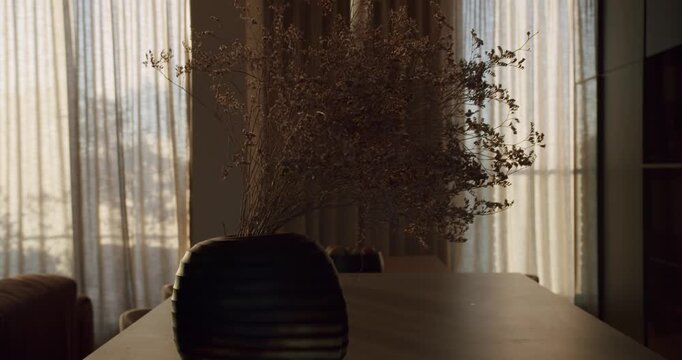 Dried flowers in a modern vase on a dining table. Sunlight streams through sheer curtains, creating a warm, inviting atmosphere. Perfect for home decor, lifestyle, and interior design projects.