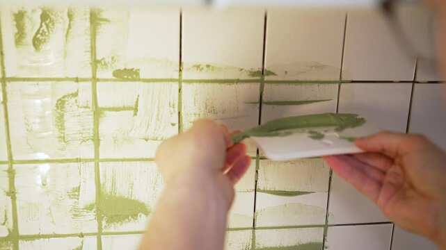 Unskilled worker's hands applying green grout to seams of white tiles on a kitchen apron with a spatula, displaying a diy home renovation project and the process of tiling