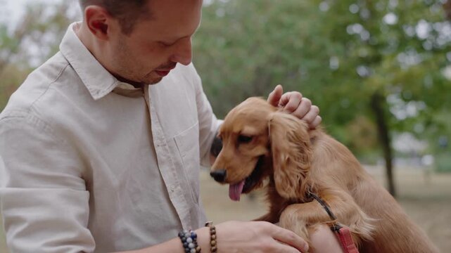 A man gently pets his adorable Cocker Spaniel puppy in a serene park setting. The connection between them is evident as the puppy enjoys the affection.The man smiles, showing love and care for his pet