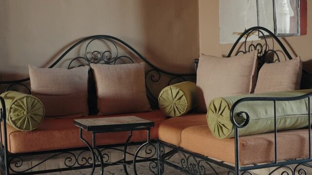 Traditional Moroccan interior design with orange and green cushions and bolsters adorning a wrought iron sofa. A small wooden table sits in front, adding to the rustic ambiance.