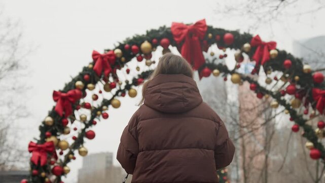 White woman under holiday arch with ornaments, brown puffer coat facing city skyline, red bows and gold baubles framing overcast urban plaza, quiet festive pause, seasonal atmosphere and wonder