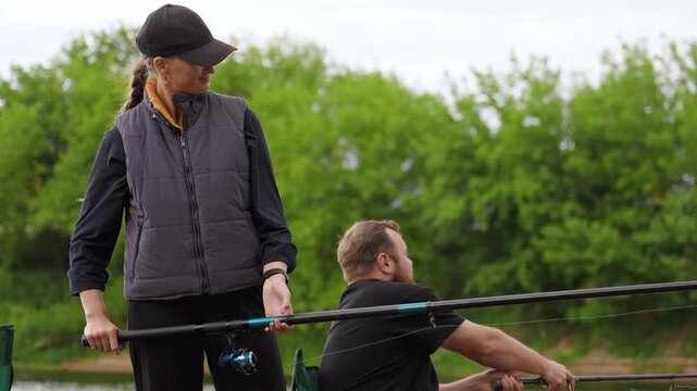 Calm couple fishing on a riverbank during an overcast day, holding rods and reels while waiting patiently for a catch. Green trees and a cloudy sky provide a serene natural backdrop