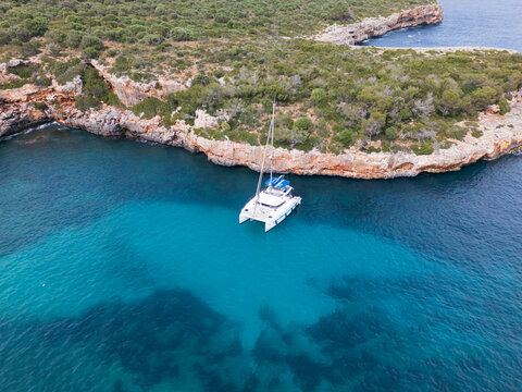 Aerial view of a white catamaran anchored in the turquoise waters of Cala Sa Nau cove surrounded by rocky cliffs and green trees in Felanitx, Balearic Islands, Spain.