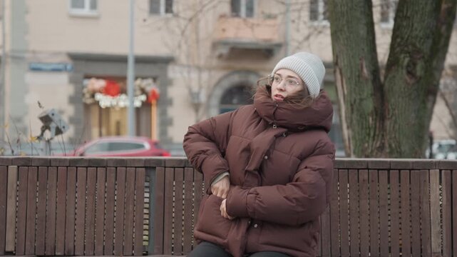 Caucasian woman calls for help on bench while rubbing hands to warm, holding smartphone and wearing thick puffer and beanie closeup frames show anxious expression behind glasses and visible breath