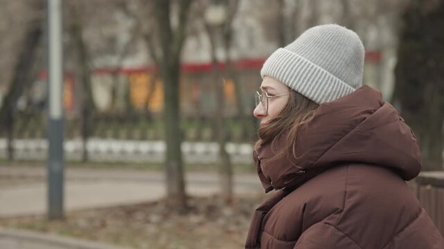 Caucasian woman commuter walking and gesturing along park path, winter layers, knit hat and glasses, expressive side profile, urban background with trees and soft lights, dynamic movement and natural