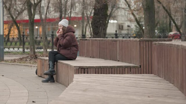 Elderly lady scrolling messages outdoors. Older lady in warm coat reviewing messages on park bench. Elderly woman calmly scrolling her phone with suitcase nearby in city park surroundings