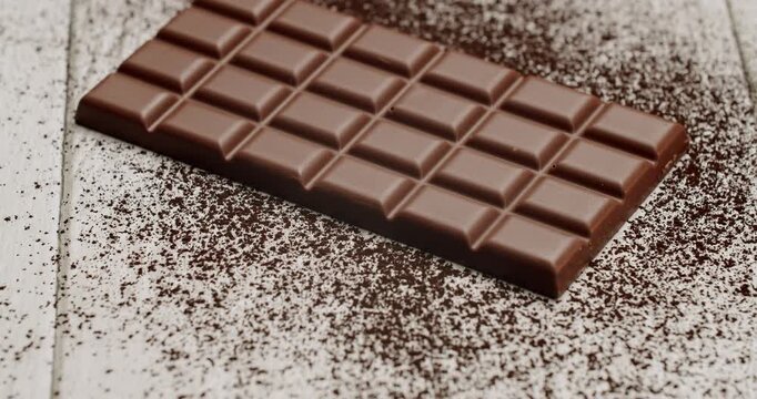 A delectable milk chocolate bar placed on powdered cocoa, creating a rich and indulgent feel. Set against a white wooden background, the scene highlights the chocolate's smooth texture.