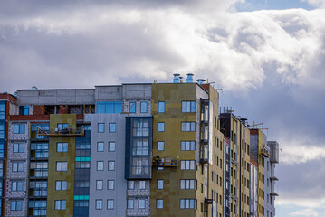 Fototapeta premium High-rise apartment building under construction, featuring multiple facades, thermal insulation, windows, and exterior finishing work in progress beneath a cloudy sky, symbolizing urban development