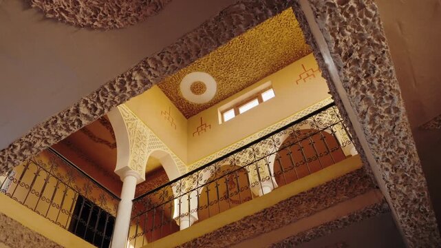 Looking up at the intricately designed ceiling and metal railing balcony of a Moroccan riad guesthouse. The warm yellow and brown tones create a cozy atmosphere, traditional Moroccan architecture.