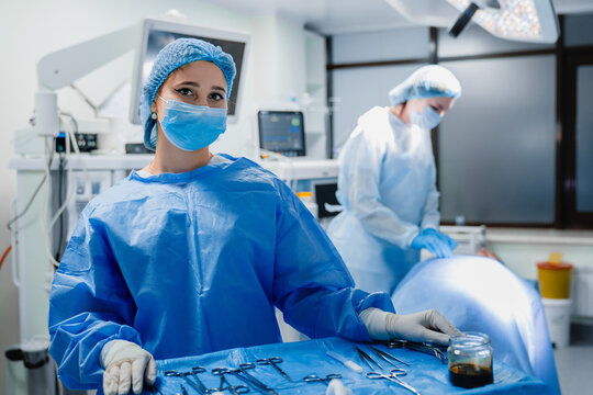 Female surgical assistant in mask standing by instrument table in operating room, sterile tools in foreground, surgeons working in blurred background, professional healthcare and surgery concept