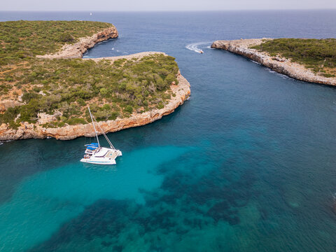 Aerial view of Cala Sa Nau with a white catamaran anchored in turquoise water and people on deck near rocky cliffs and green vegetation in Felanitx, Balearic Islands, Spain.