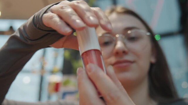 Closeup woman opening lipgloss bottle, greenhouse backdrop with string lights and plant bokeh, hands unscrewing white cap to reveal translucent pink serum, delicate applicator visible, casual glasses