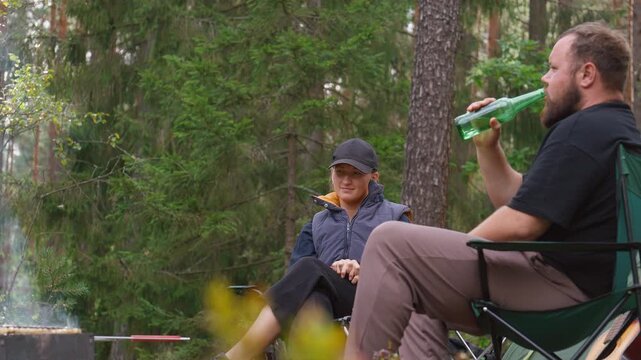 Young man and woman sitting on folding chairs in pine forest, jingling beer bottles and enjoying barbecue picnic while camping, with grilled food on small charcoal barbecue