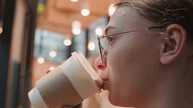 Young white woman sipping coffee by window in quiet cafe, slow mindful sip from paper cup, steam and foam catch warm lights, contemplative profile with glasses, productive pause during research