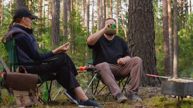 Happy adult couple enjoying a relaxing picnic in the middle of a sunny pine forest, sitting on camping chairs, drinking beer, and cooking food on a barbecue grill during a weekend getaway