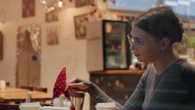 Handmade holiday crafting with red gnome, woman shaping felt ornament at cafe table, warm lights and pastry counter in background, focused gentle smile and seasonal cozy atmosphere