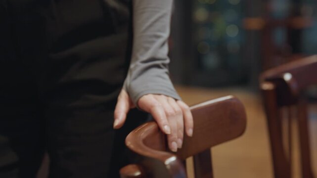 Asian server preparing wooden chair with gentle hand, closeup on fingers and backrest, dark apron and grey sleeve, warm wooden floor and blurred dining tables in background, focused routine