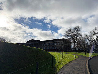 Obraz premium Sunlit neoclassical National Galleries Scotland, Modern One building on a grassy hill with winding path and bare trees in Edinburgh, Scotland, UK under a dramatic cloudy sky