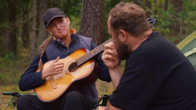 Young smiling woman playing an acoustic guitar and singing songs for a bearded man while sitting in camping chairs in the woods. Happy friends enjoying their vacation and music together in nature