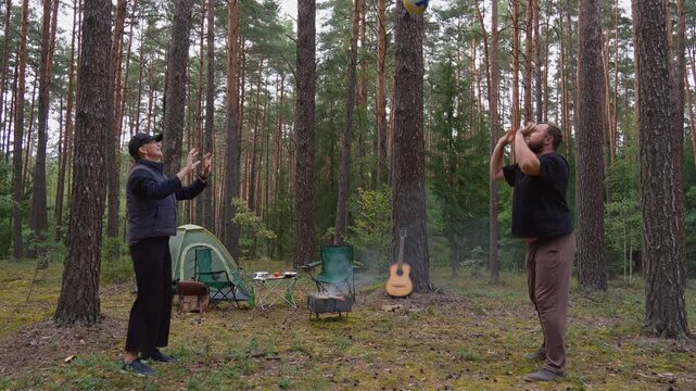 A young couple woman and a man play volleyball in a forest clearing while camping, a tent, a fire and a guitar can be seen in the background, which contributes to an active family vacation