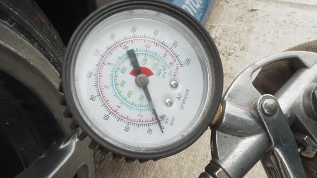 Closeup pressure gauge on tire showing needle in green zone, metal valve chuck and hose visible, chrome fittings, concrete workshop background, precise measurement for automotive maintenance