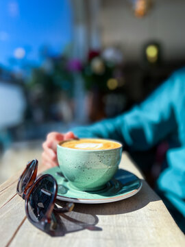 A hand holds a fresh cup of cappuccino on a wooden table, with stylish sunglasses placed nearby, creating a relaxed morning or vacation vibe.