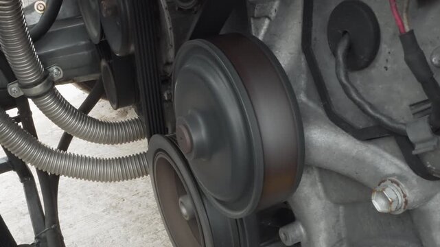 Close up spinning drive pulley and belt, rust toned pulley wheel rotating against serpentine belt, braided hoses and visible tensioner, surface wear and grime indicating service need, motion blur