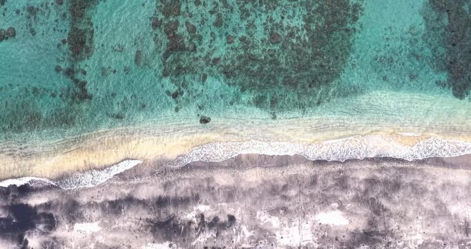 Overhead aerial drone shot of turquoise coral reef waters meeting the volcanic black sand shoreline in Amed, Bali, Indonesia.