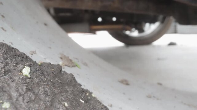 Low angle undercarriage dirt closeup, parking garage floor shows mud pile and discarded paper tube, scattered green leaf fragments, exposed wheel and chassis, gritty texture, mechanic inspection