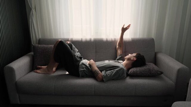 Wide sofa shot caucasian man reaching toward sheer curtain, fingers playing with light, knees bent and feet bare, contemplative gesture and quiet longing, neutral interior