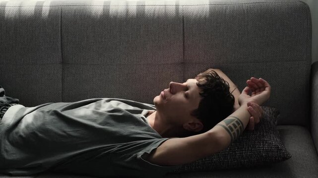 Sunlit couch closeup caucasian man lying on grey sofa, tracing ceiling with fingers, stretching and shielding eyes, silent daydreaming with visible forearm tattoo, soft pillow
