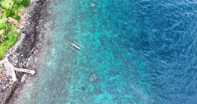 Top-down aerial drone view of the rocky coastline and vibrant coral reef waters in Amed, Bali, Indonesia with a traditional Balinese jukung boat floating near the shore.