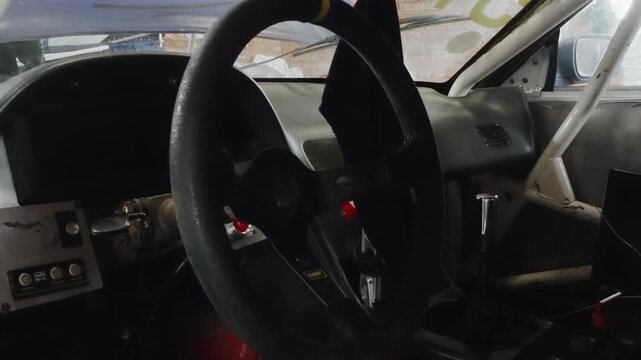 Rally cockpit steering wheel closeup rugged dashboard showing shifter and harness, textured leather grip, switch panel, roll cage frame, ambient garage light, ready for race launch
