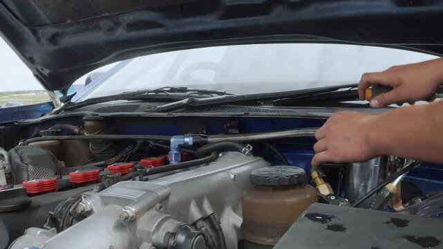 Hands repairing car engine under hood, closeup of screwdriver adjusting intake manifold, red ignition coils, oil stains on metal, technician performing manual tune, outdoor garage lighting, focused
