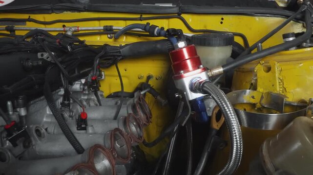 Yellow engine bay with braided hoses and red regulator, closeup of exposed intake manifold, ignition coils and metallic valve covers, workshop lighting highlighting fittings and steel braided lines,