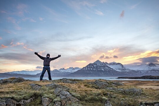 Hiker with outstretched hands standing at the fjord Berufj&ouml;r&eth;ur, sunset over snow-covered mountains, Austurland, East Iceland, Iceland