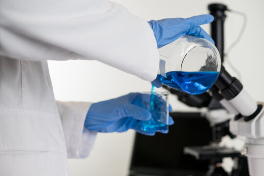 A scientist transfers a blue liquid from a larger flask into a beaker, representing the titration or mixing process in Hantavirus research.