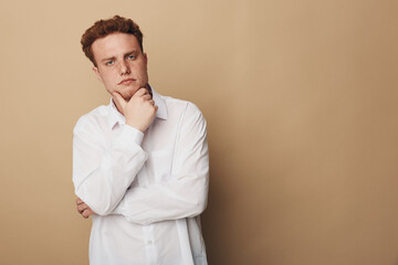 Thoughtful young man in white shirt stands against beige background with hand on chin. Concept of contemplation, decision making, and casual style for lifestyle and portrait. © SHOTPRIME STUDIO