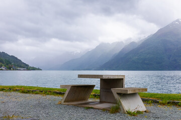 Quiet escape by Hardangerfjord with concrete table facing calm water and layered mountains with rain clouds in western Norway. Contemplative travel, Scandinavian outdoors and peaceful nature themes. © Nimbus Works