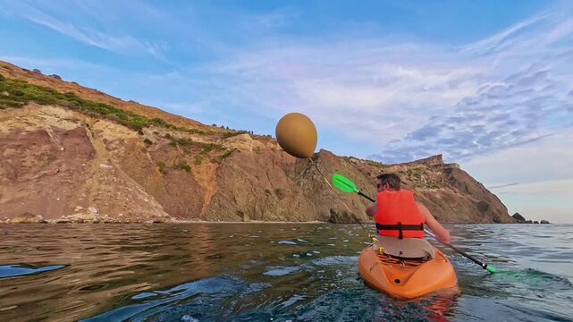 Kayak paddling ocean man in orange life jacket paddles small boat past tall rocky cliffs on sunny day