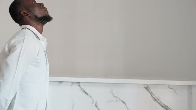 Black man standing beside marble counter, looking upward and exhaling, adjusting collar with calm posture, quiet acceptance and resolve after late date, modern interior and steady composure.