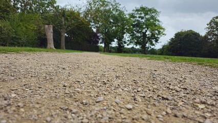 Fototapeta premium Low angle view of a wide gravel pathway leading through a green park with trees under a cloudy sky, England, UK