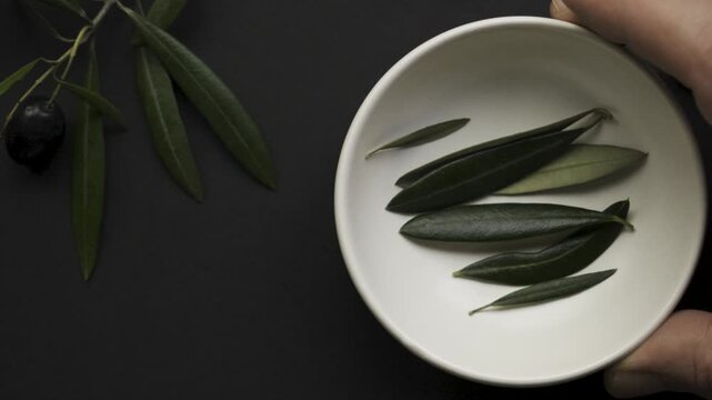 Hand_adjusting_white_bowl_with_olive_leaves_on_black_background_slomo_4k