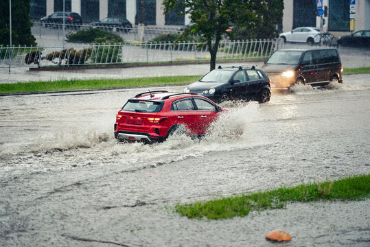 Red hatchback creates massive white splash while driving through deep flood water on city street during heavy seasonal downpour with other cars visible in the background. Cars navigating flooded road