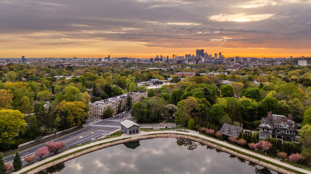 Aerial view of Brookline Reservoir surrounded by lush spring cherry trees and residential houses with the Boston skyline in the distance at sunrise. Brookline, Massachusetts, United States.